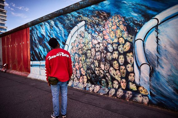 Image from the East Side Gallery by artist Kani Alavi titled It´s Happened In November. A man in a red top stands facing away from the camera admiring the painting. The painting is of many faces rushing through a wall with a blue, sea-like background