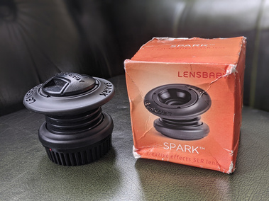 The Lensbaby Spark 1.0 next to it's box. The box is orange and the lens is black, it has a lens cap on and plastic squeezable bellows so it is able to be manually positioned to chose focus points