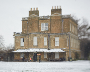 Clissold Park House, a grand house with many windows and chimneys. There is snow on the ground and the edges of the frame are soft - a creative effect of the Lensbaby Spark. There is a person walking in the distance with a red and orange umbrella
