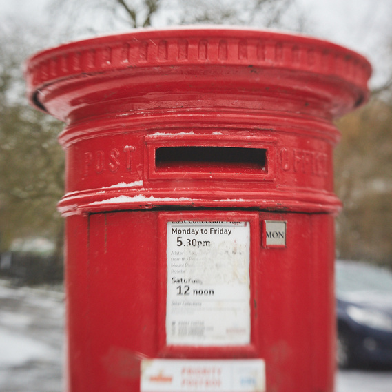 Square image of a red post box. The background has soft focus with beige and white tones, hinting at a quiet London street covered in snow