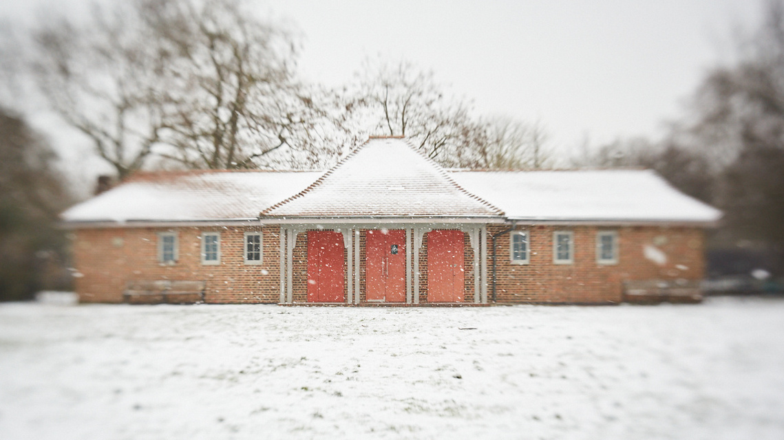 Clissold Park Education Centre, a brick pavilion in a snow-covered park. There are pale red doors and wooden beams in the centre of the building. 