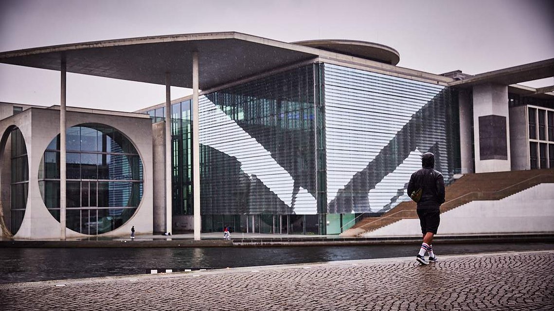 A man dressed in black walks in the rain past the Marie-Elisabeth-Lüders-Haus. It is a large glass building with light reflections across the Spree River