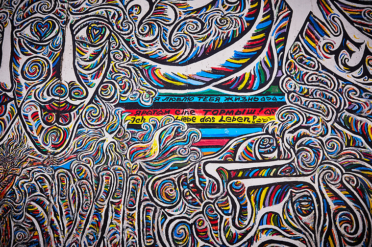 Straight on view of a multicolour mural at the East Side Gallery by artist Schamil Gimajew titled Worlds People. Within the mural are stars, faces and flowers outlined in black