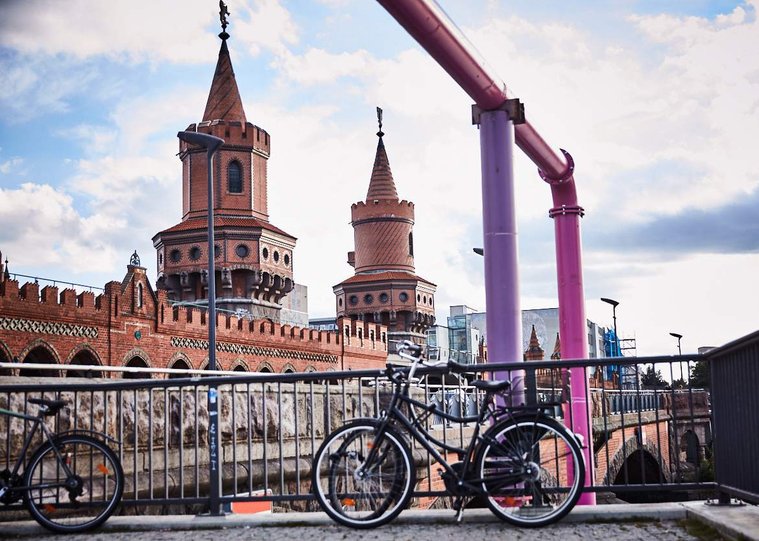 Turrets of the Oberbaum Bridge, a double-deck bridge running across the River Spree. Bicycles can be seen against the railings in the foreground and pink metal tubes frame the image