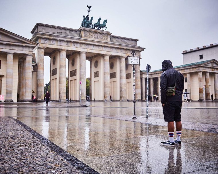A man dressed in black stands in front of the Brandenburg Gate. It is raining and he has his hood up and face down, away from the camera