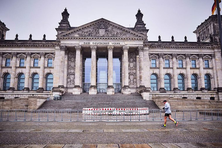 A man jogs past the Reichstag building. It is raining and looks cold. The building has tall stone columns and a railing outside, preventing crowds during the COVID-19 pandemic. The scene is empty of people except for the one lone jogger