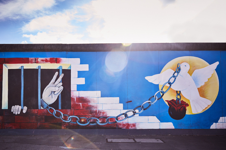 Image of the Dove of peace, carrying a chain in its mouth which is attached to a hand with a peace sign. The hand is in jail.  From the East Side Gallery by artist Andrej Smolák. The sun shines through the lens creating flare. 