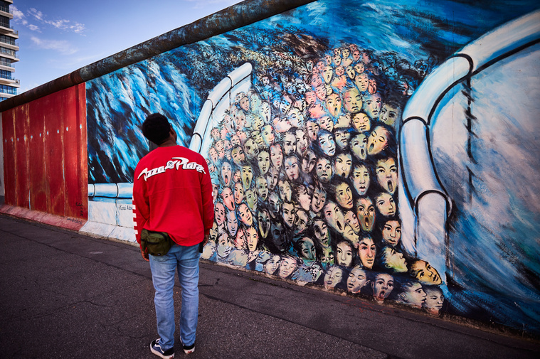 Image from the East Side Gallery by artist Kani Alavi titled It ́s Happened In November. A man in a red top stands facing away from the camera admiring the painting. The painting is of many faces rushing through a wall with a blue, sea-like background