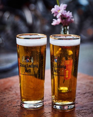 Two pints of beer sit fizzing on a wooden table. They look crisp, fresh and inviting. In the background is a lilac coloured flower