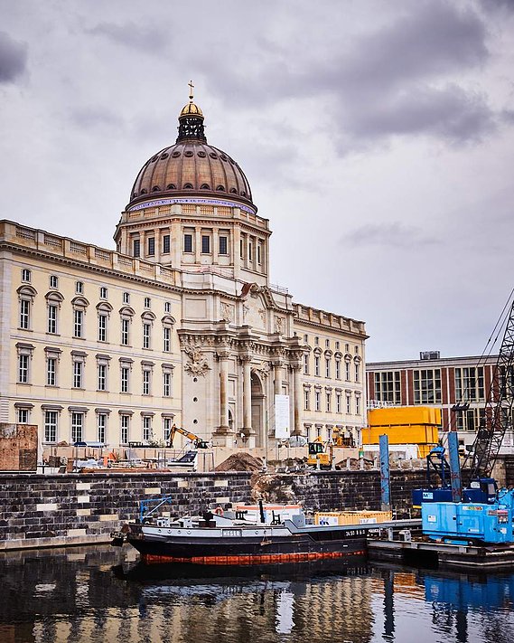 View of Berlin Palace Humboldt Forum, a grand yellow bulding with many windows. There is a gold cross on the top of a bronze dome in the centre. In the foreground you can see boats harboured in the River Spree