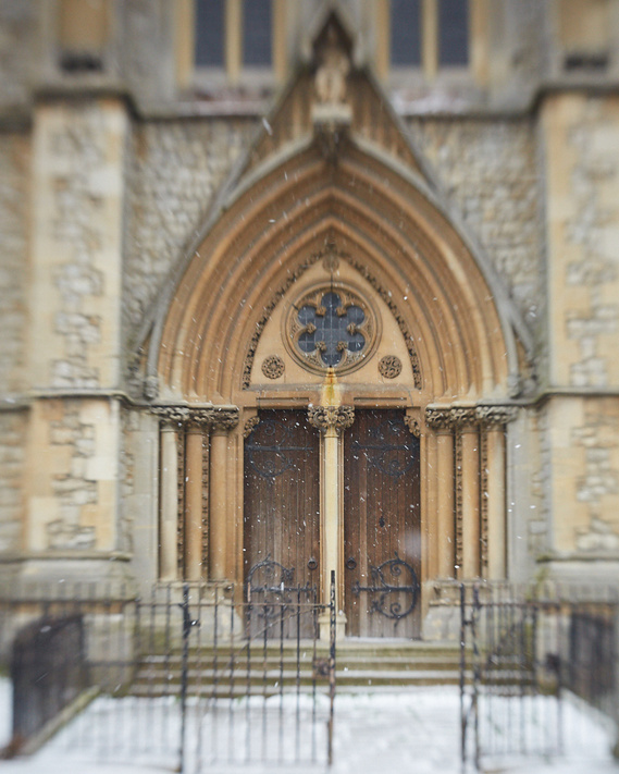 Brown church doors and an arched stone entrance to St Marys Church. There is snow visible on the ground. The edges of the image have a shallow depth of field, meaning they have soft focus.