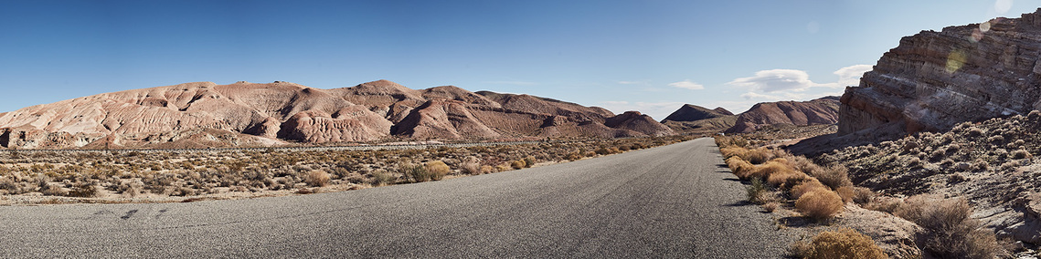 A panoramic view of red rock canyon. In the foreground is a deserted road, leading into rocky mountains in the distance. The sky is blue and the sun is beating down