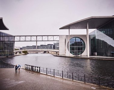 View of Marie-Elisabeth-Lüders-Haus. It is a large glass building on the Spree River. Two people can be seen walking away from the camera in the distance, heads down and hoods up to shield from the rain