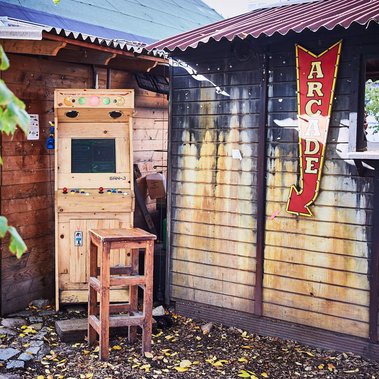 Wooden arcade joy stick game at Holzmarkt 25 with a wooden stool in front of it. There is a red and yellow vintage looking sign next to it, saying 'arcade'