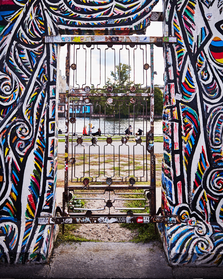 Artwork at the East Side Gallery in Berlin by artist Schamil Gimajew titled Worlds People. A rusted metal gate frames a river can be seen with people sitting on the grass in front. The artwork frames the old gate