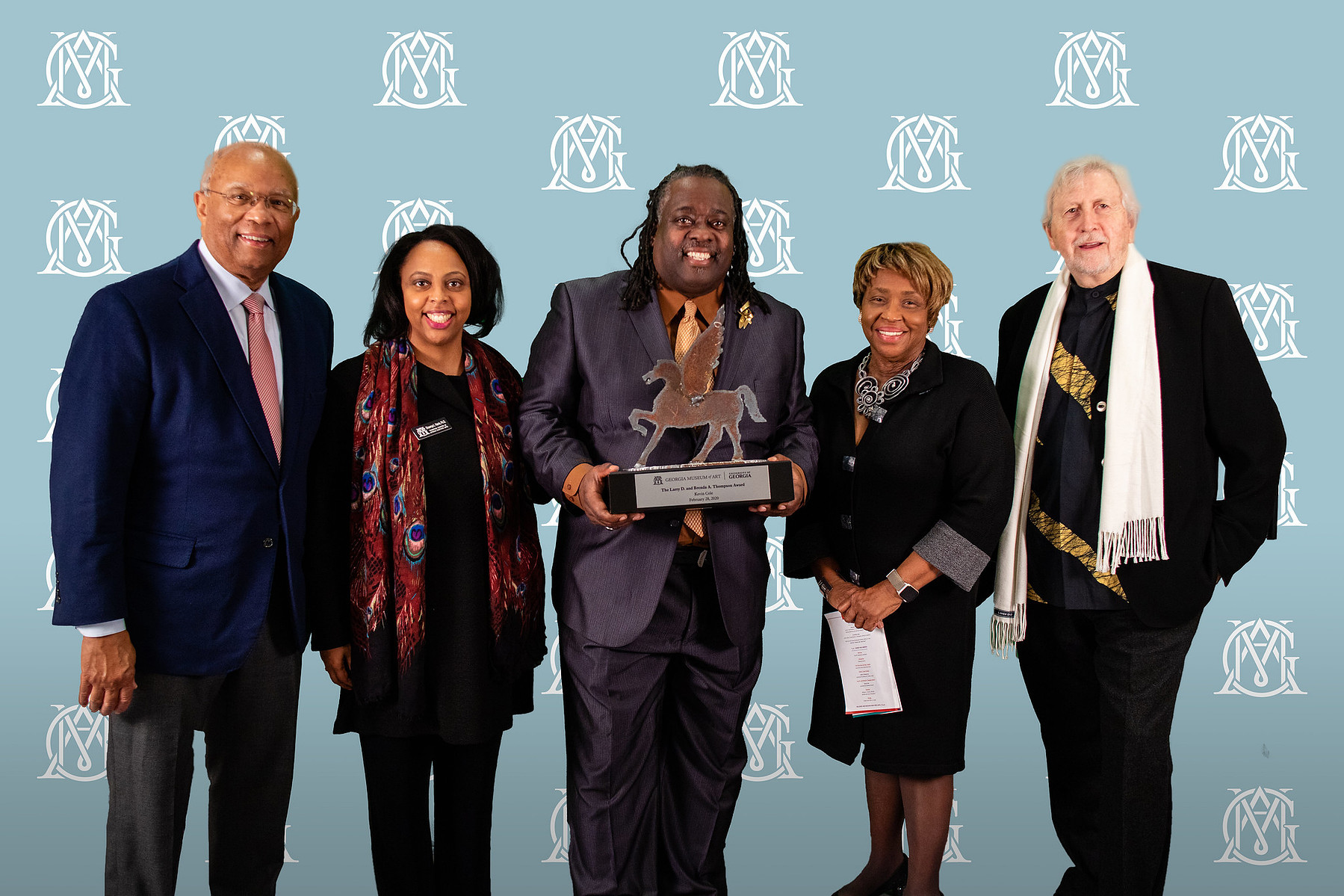 Brenda and Larry Thompson Award - Georgia Museum of Art- Five People in Professional Wear Posing for a Picture With a Man in the Middle with an Award in His Hand. The Background of The Picture is Pale Blue with a White Logo.