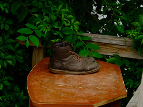 A pair of old leather work boots sits on a small table in front of green bushes. 