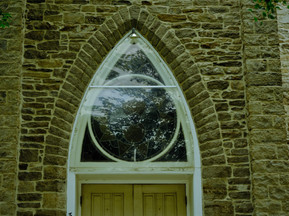 A pointy window reflects a cloudy sky.  It sits above a sturdy wooden door to a grey stone church in Kemptville.