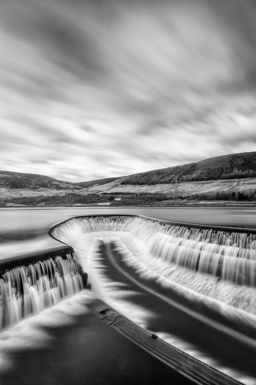 Torside reservoir in the North of the peak district with water cascading over the edge of the waterway with clouds moving in the distance due to a long exposure