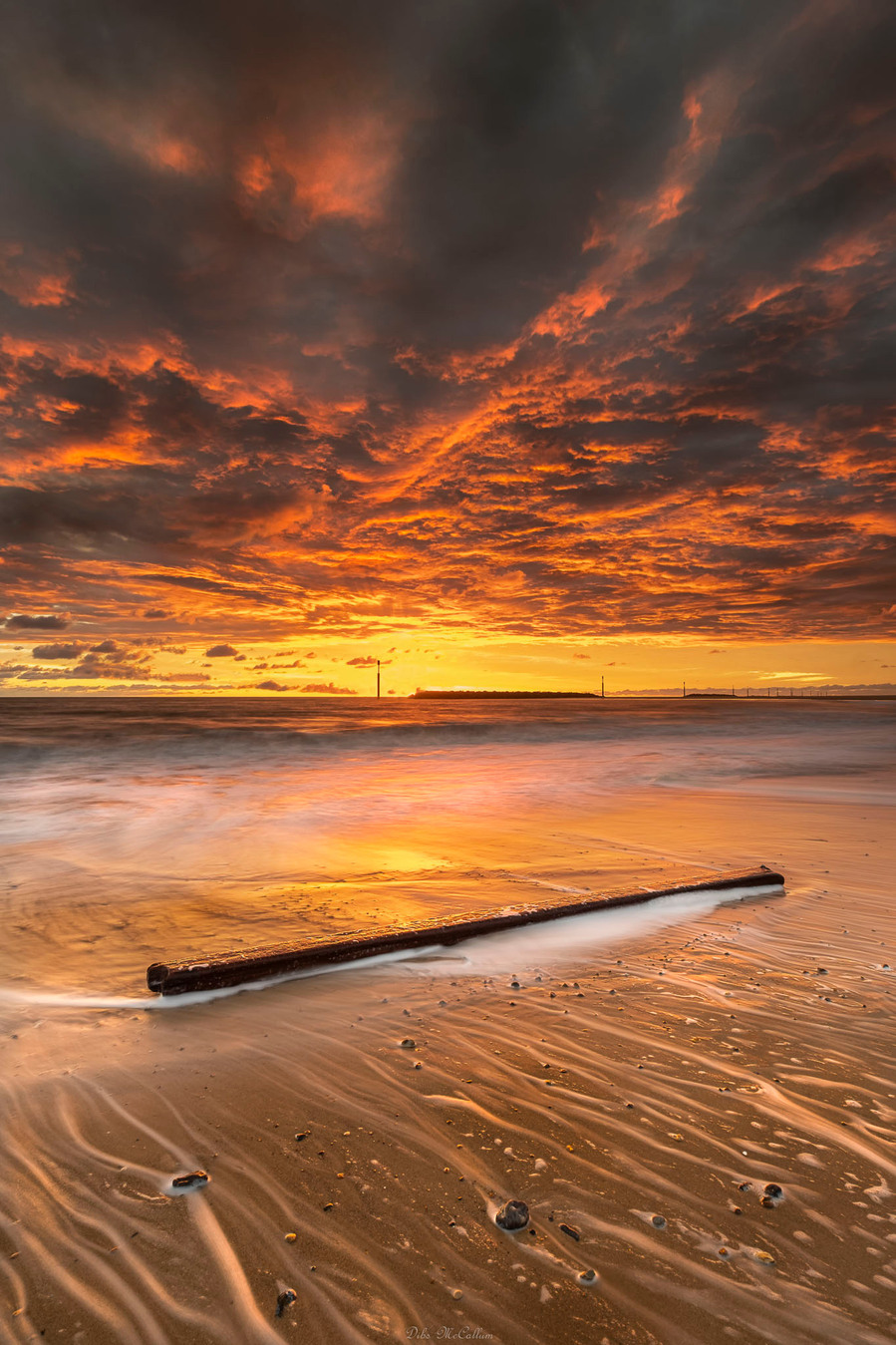 Sunrise at Sea Palling in Norfolk with a firey red sky and textured clouds and some driftwood on the beach with waves splashing on it