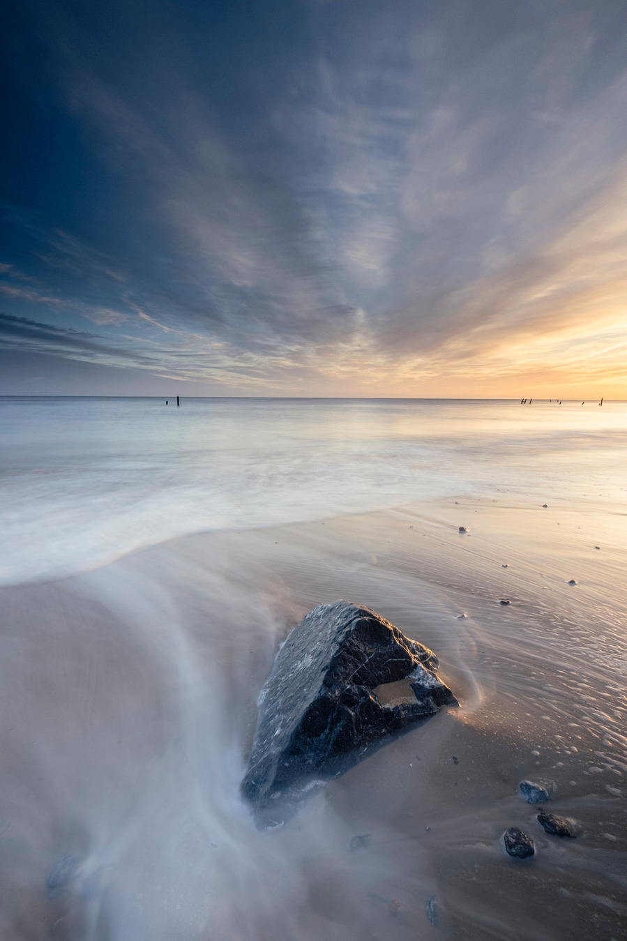 A singular rock laying on the beach at Happisburgh in Norfolk with the golden light of sunrise mixing in with the blue of the blue hour as waves run past the rock creating white streaking on the sand