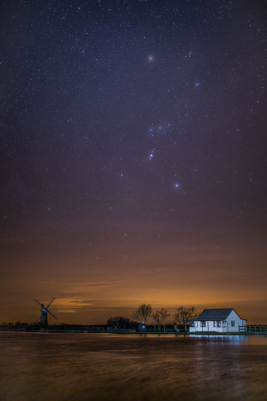 Thurne mill with st Bennets drainage mill and a white riverside house and the Orion star constellation above it with a glow from the nearby light pollution in the sky and also on the river Thurne that is part of the Norfolk broads national park