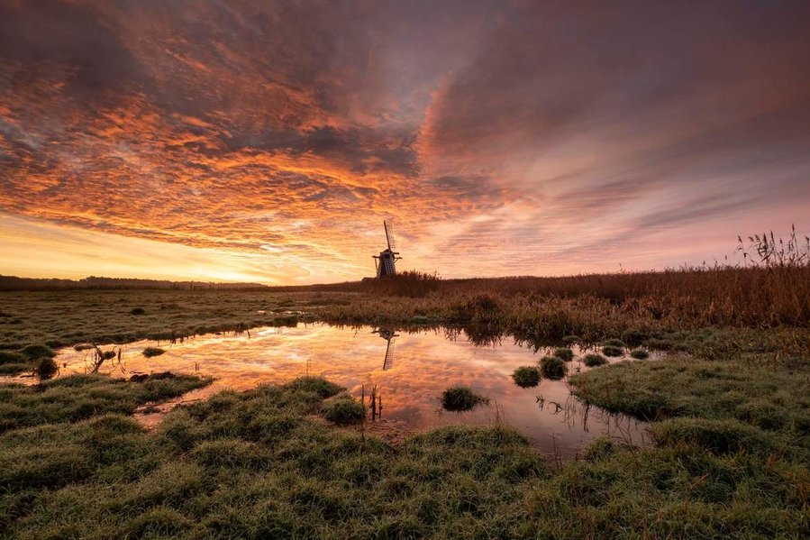 Herringfleet mill that is part of the Norfolk Broads at sunrise with a firey red sky and clouds, with a reflection of the windmill and the sky in a flooded part of a field that has a little bit of green grass and reeds near it