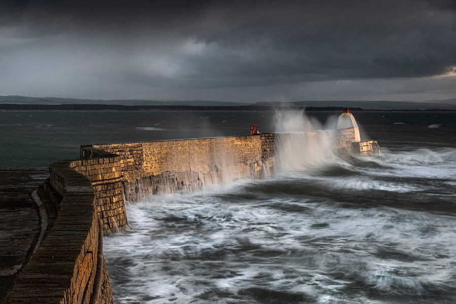Waves crashing on the Burghead harbour wall in Moray during a storm with the whites of the sea water visible and the sunlight illuminating the brick work on the wall