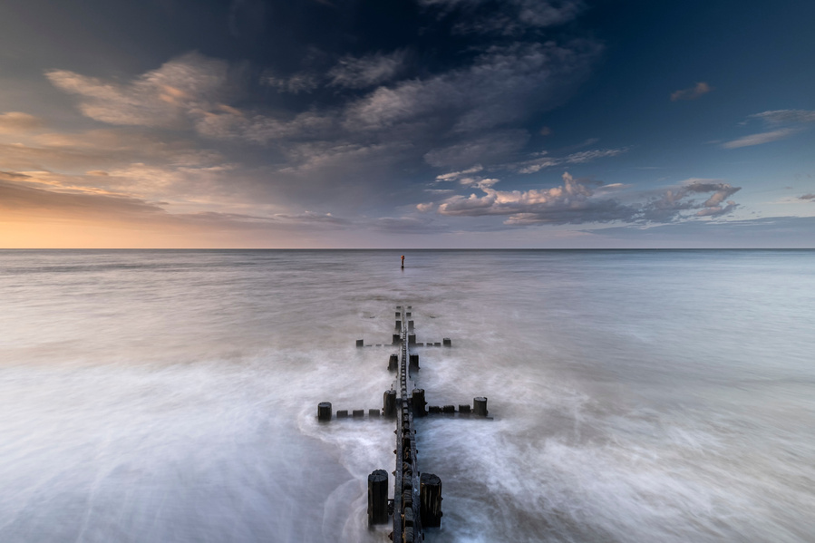 The sea defences running straight out into the North sea at the bottom of the cliffs at Mundesley in Norfolk at sunset with a mix of golden light and blue light in the sky with clouds catching the light as the sea is smoothed out with a long exposure