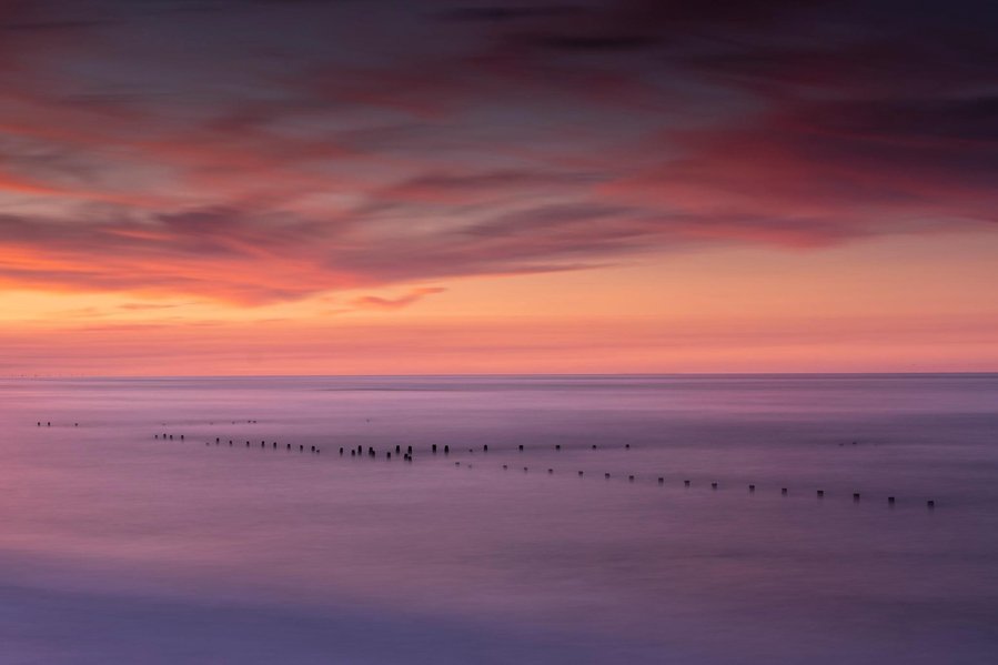 An amazing red sky sunset of the beach at Happisburgh in Norfolk with the wooden sea defences poking out of the still water of the North sea as the sunlight reflects onto the smoothed out sea created by a long exposure