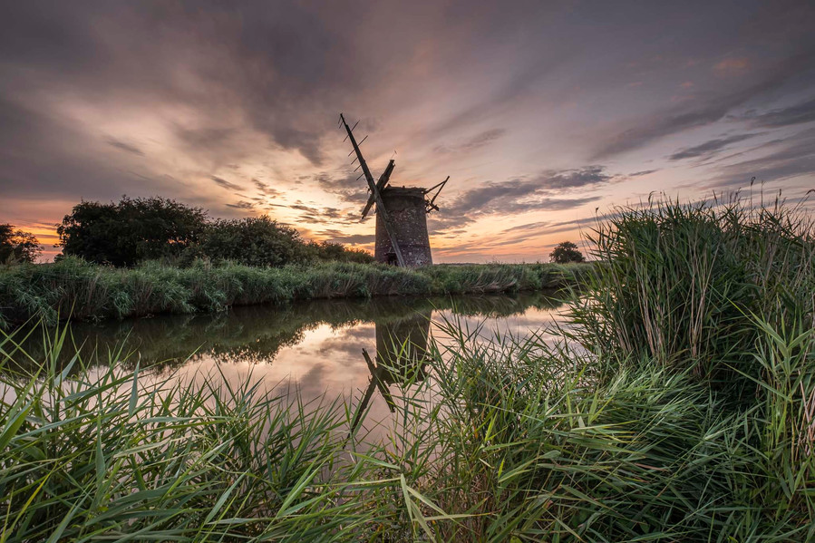 Norfolk broads windmill at sunset with green reads around it and a reflection on the water