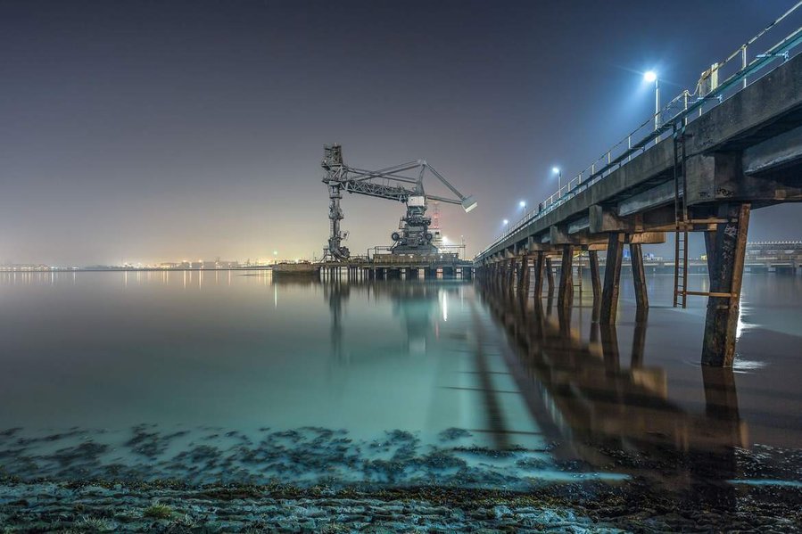 The derelict and unused Thurrock coal lifter machinery on the river Thames with a blue and green hue from the floodlights and a reflection on the water