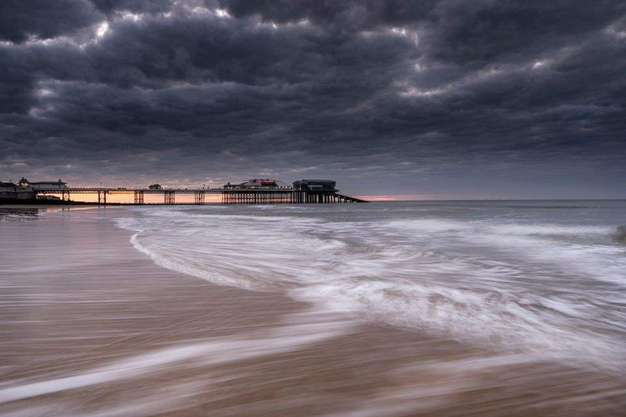 Cromer pier at low tide at sunset with the waves moving on the golden sand beach and a moody sky