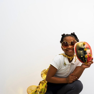 a smiling Black girl in a gold cape crouches holding a painted mask