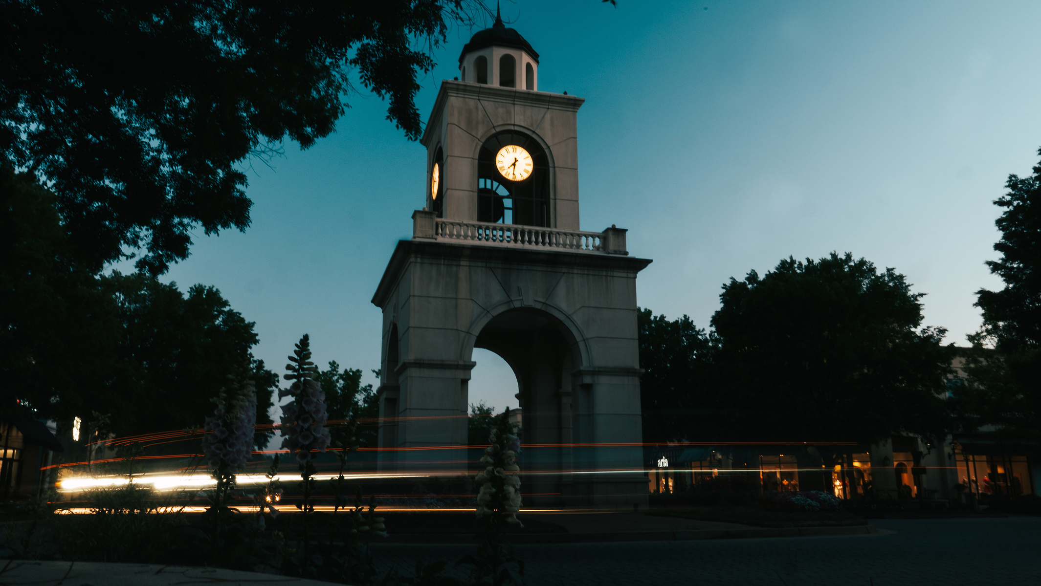 Headlights circling the clock tower at the center of a shopping mall at dusk.
