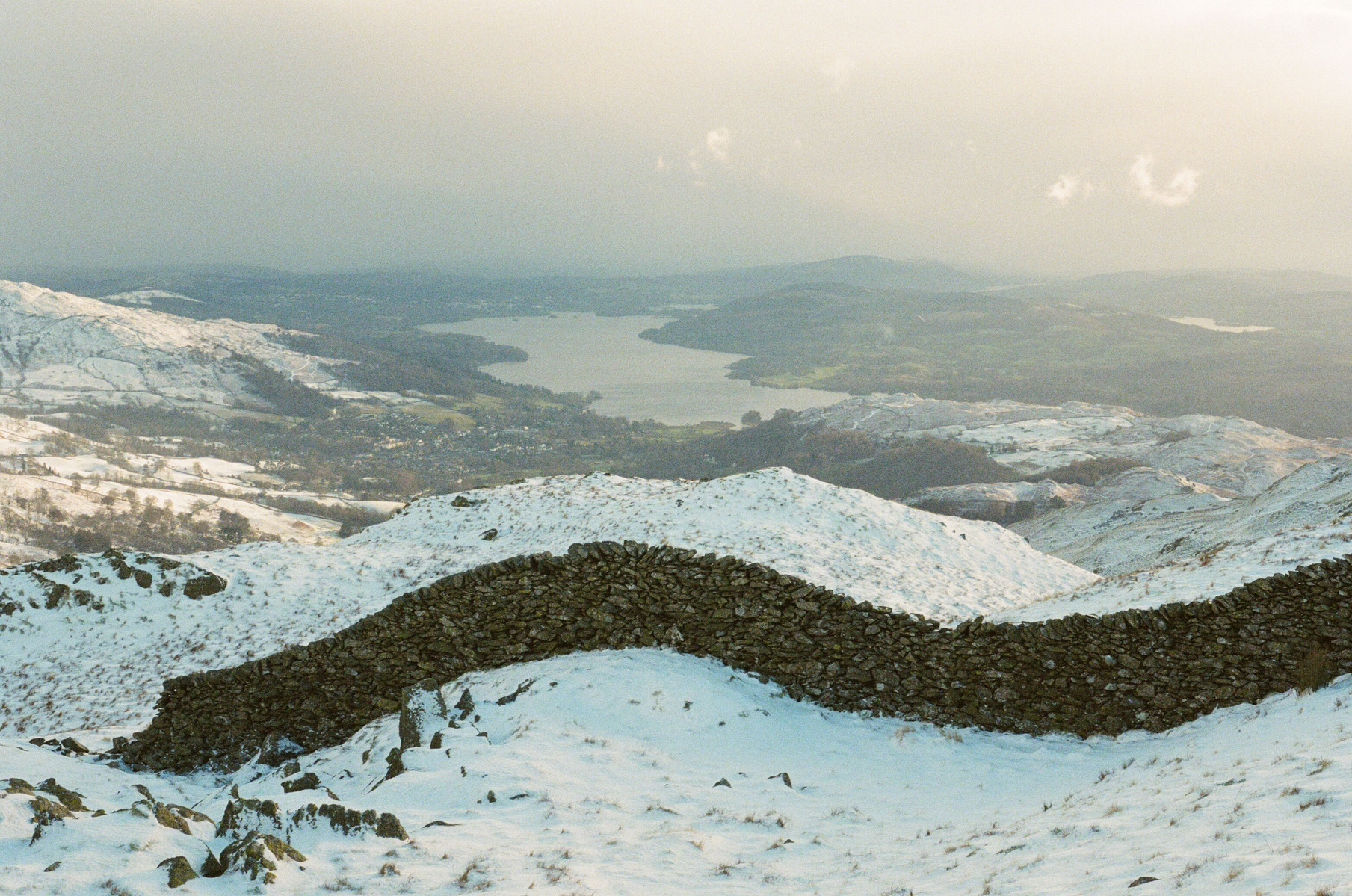 A dry stone wall snakes over a snowy fell overlooking Windermere in the Lake District