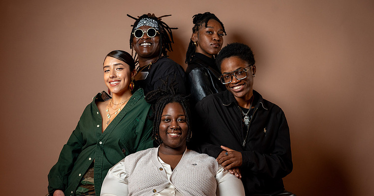 This image features five, Black and Brown people who are members of a writers group, The Write it Down Collective. Two people stand in the back, two are seated on the arm rests, and one sits front center. They are all smiling and looking at the camera.