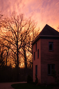 Bare tree branches and the side of a building are starkly silhouetted against an intense sunset sky.