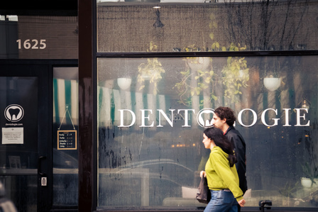 A man and a woman are walking by a storefront with the words "Dentologie" painted on the window