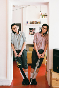 Portrait of a twin holding an umbrella each and leaning sideways on a doorway. The photograph is shot using analog film.