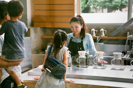 A girl leaning over the counter as she is being served by a smiling female staff of Blue Bottle Coffee Japan.