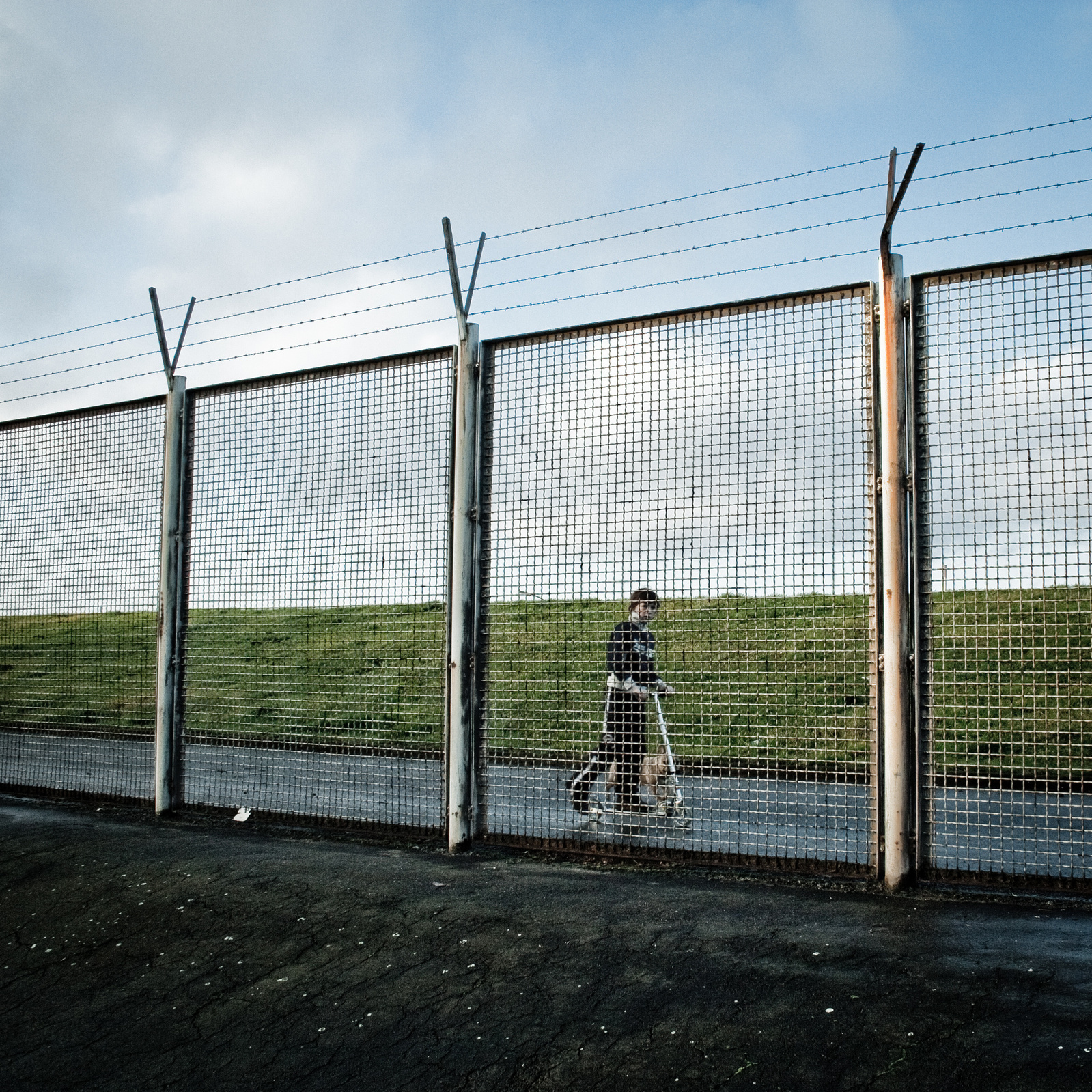 Lonely little boy with a scooter behind a large fence.