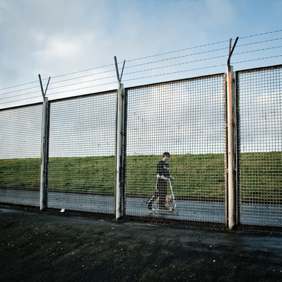 Lonely little boy with a scooter behind a large fence.