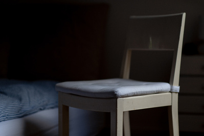 Dark and moody still life photo of a white chair next to a bed.
