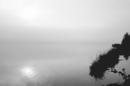 Calm water with sun reflection through the fog, in distance a man fishing from grassy strand