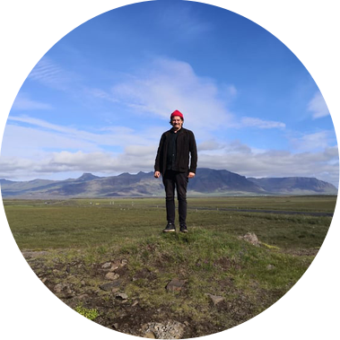 Man with red hat standing on a small hill in open landscape with mountains in distance.