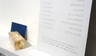 Detail photo of an installation of contemporary art. The photo depicts a ledge holding three objects: two prints and a clear box of gold leaf.