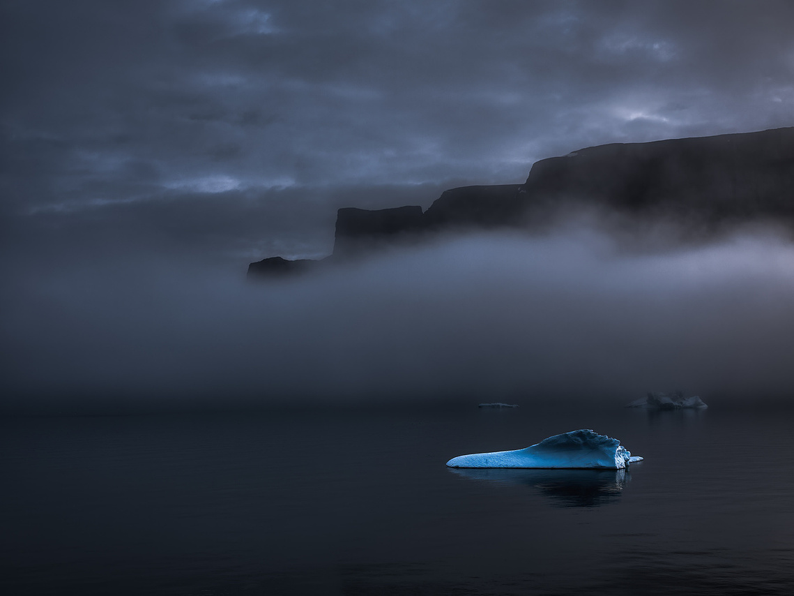 A solitary blue iceberg floating on dark, still water beneath fog-covered cliffs and a heavy overcast sky.