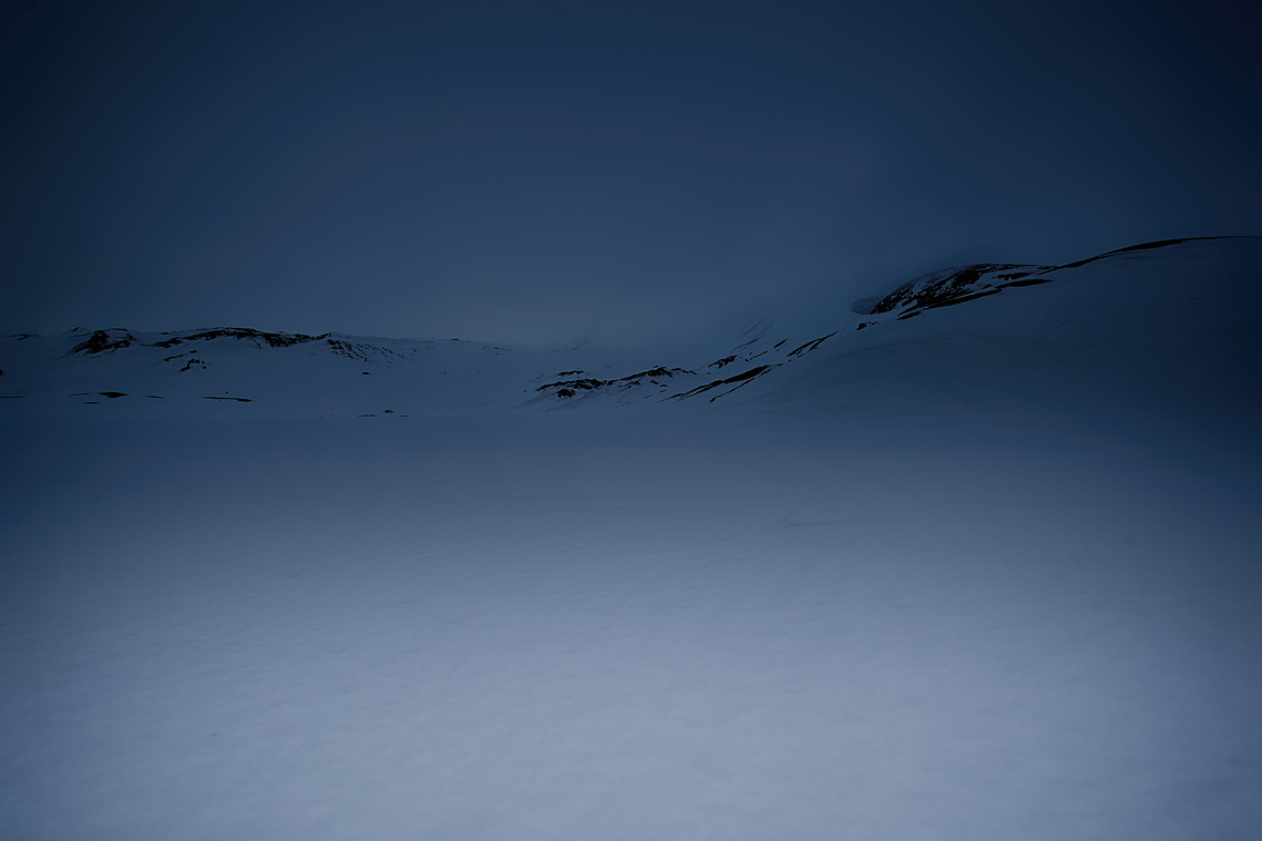 Minimal polar landscape with a vast snow-covered plain and low dark ridges beneath a deep blue twilight sky.