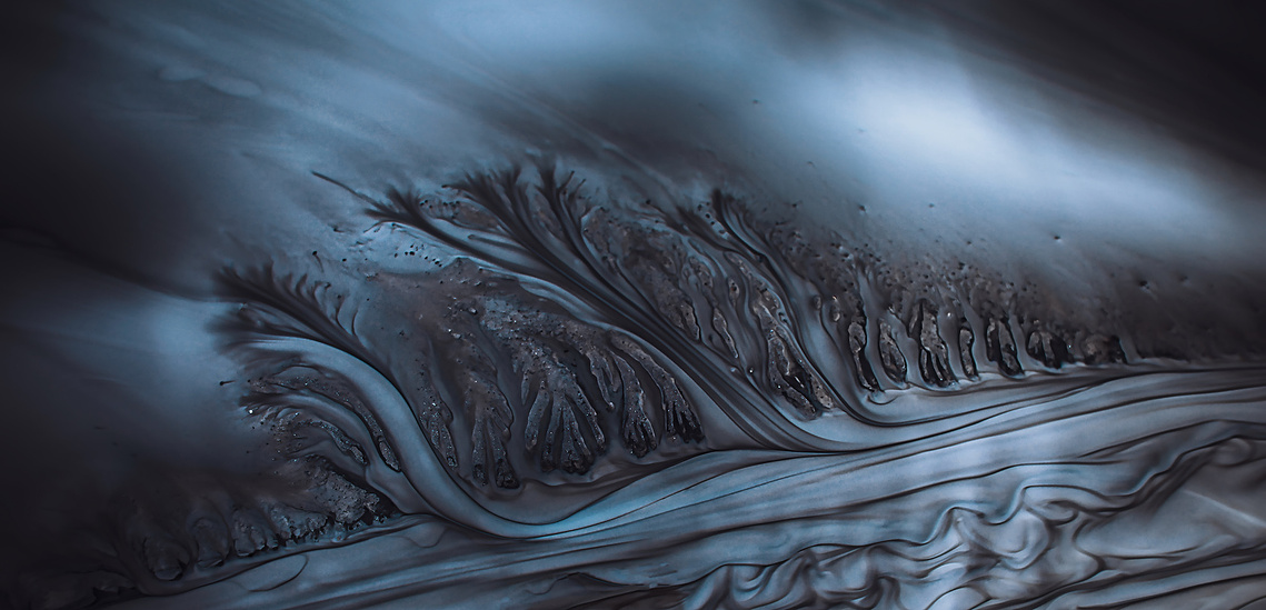 Abstract close-up of dark mineral sediments forming branching, fluid patterns beneath a band of cold blue light.
